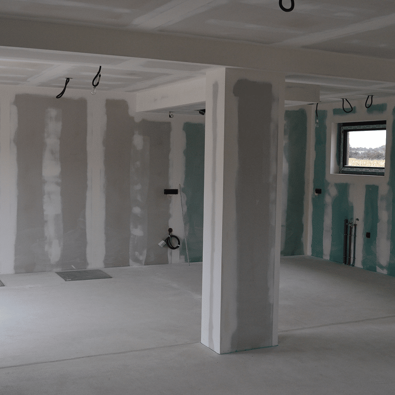Intérieur de maison en construction: murs en placo enduit, plafond, sol béton brut, colonne et fenêtre visible. Préparation avant finitions.