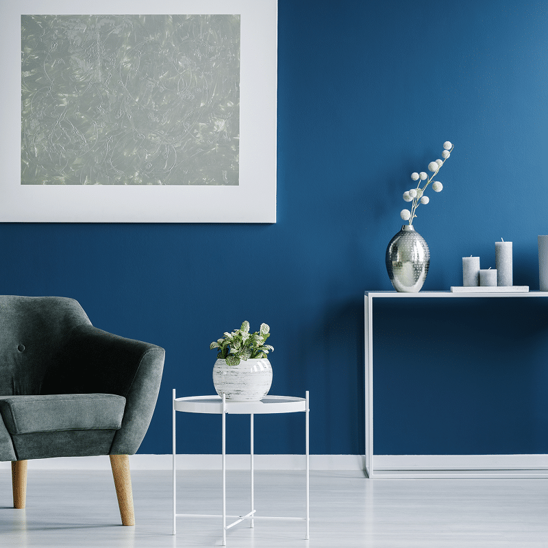 Salon moderne aux murs bleu marine, fauteuil vert foncé, table basse blanche avec plante, console avec vase argenté et bougies grises.