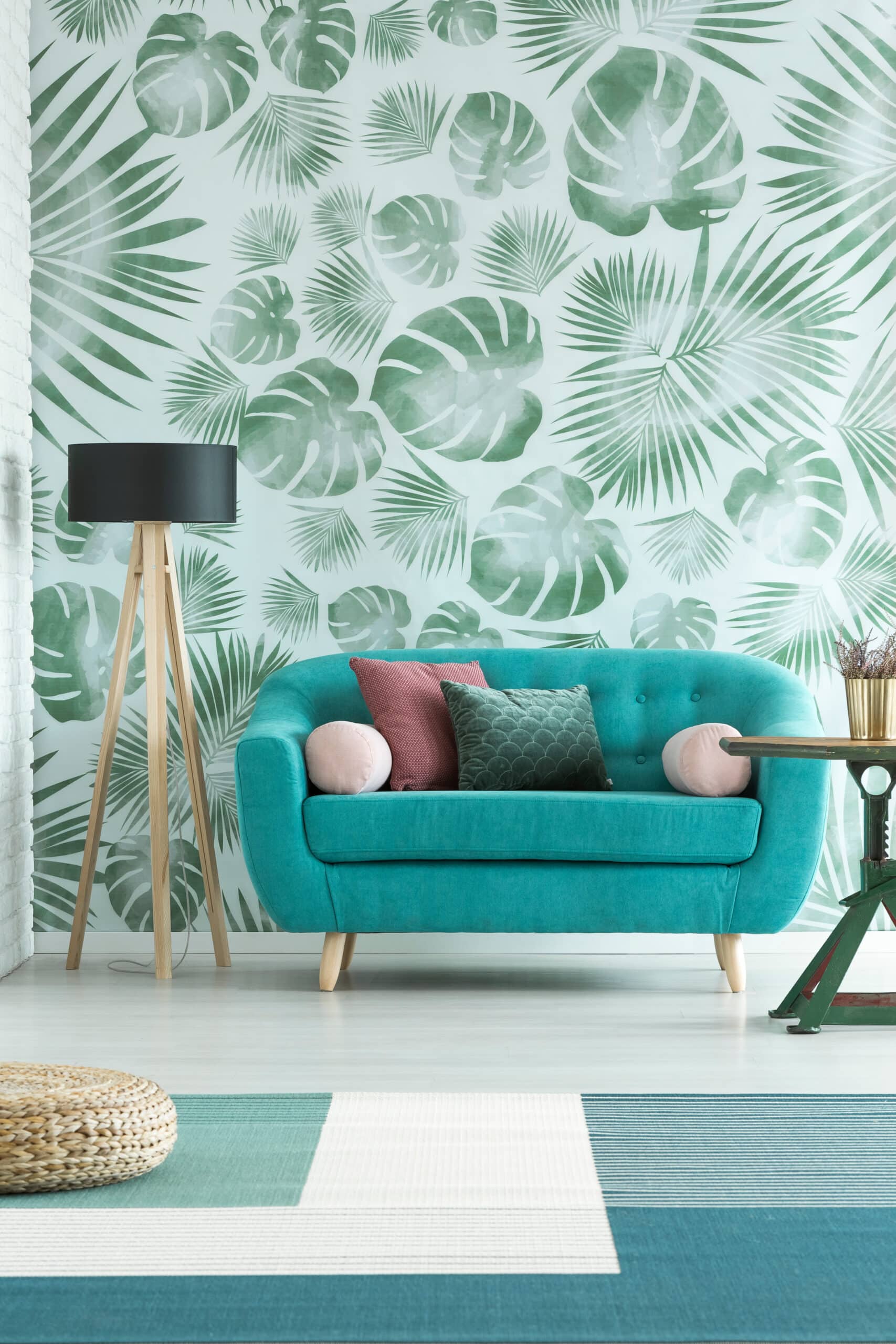 Salon moderne avec canapé turquoise, coussins colorés, lampadaire trépied bois. Mur tapissé de feuilles tropicales vertes, pouf tressé sur tapis rayé.